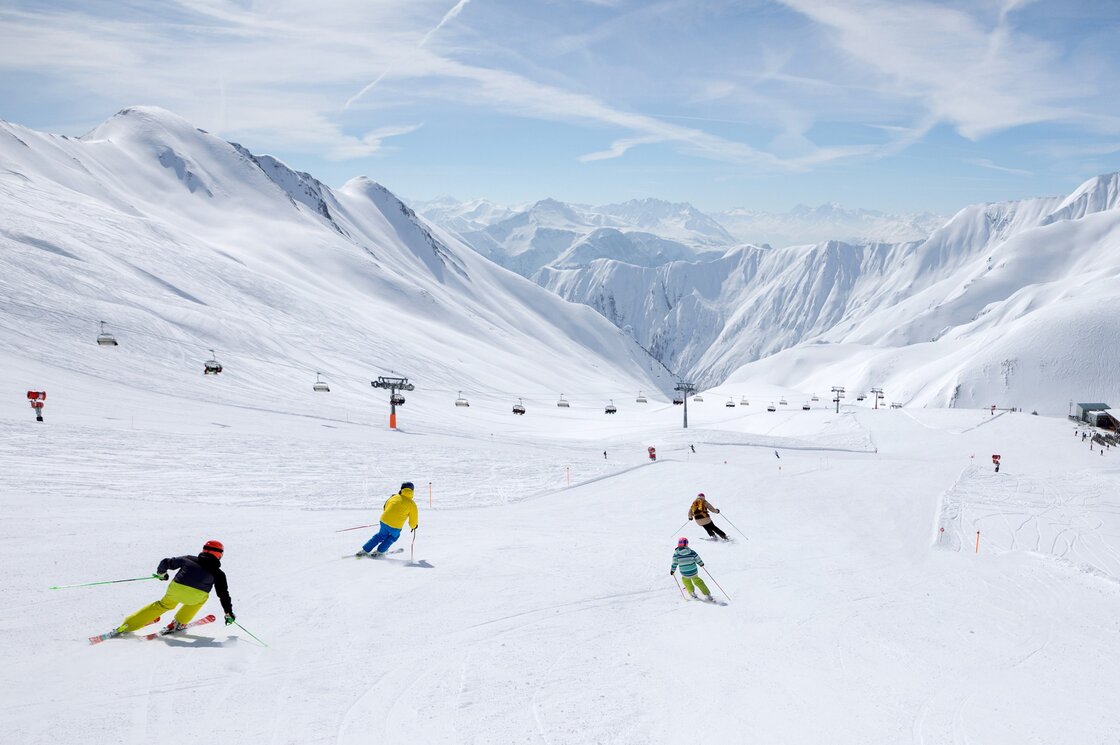 Mehrere Skifahrer fahren auf einer breiten Piste im verschneiten Skigebiet zwischen hohen Bergen.