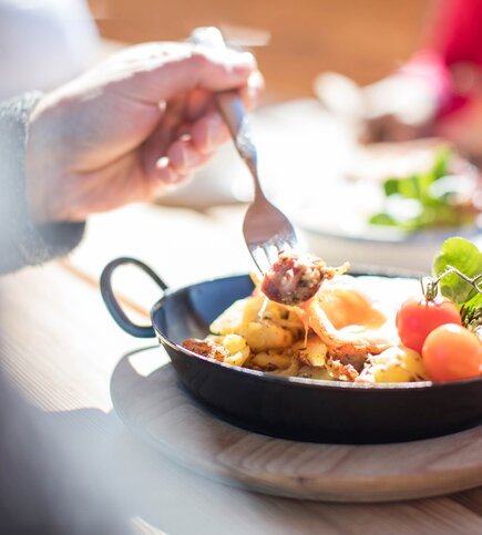 Eine Hand hält eine Gabel über einer kleinen Pfanne mit Frühstück und frischen Tomaten.