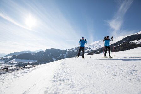 Zwei Langläufer stehen auf einer verschneiten Hügellandschaft mit Bergpanorama unter blauem Himmel.