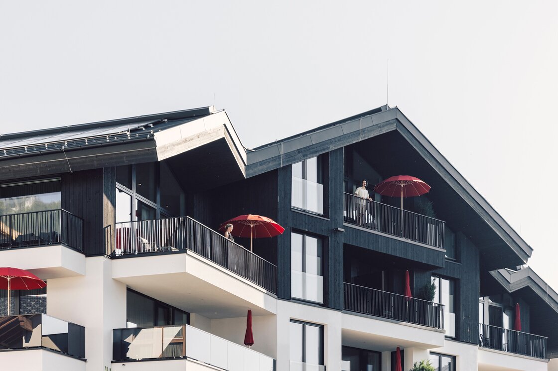 Das moderne Apartmentgebäude zeigt dunkle Holzfassaden, weiße Balkone und rote Sonnenschirme.