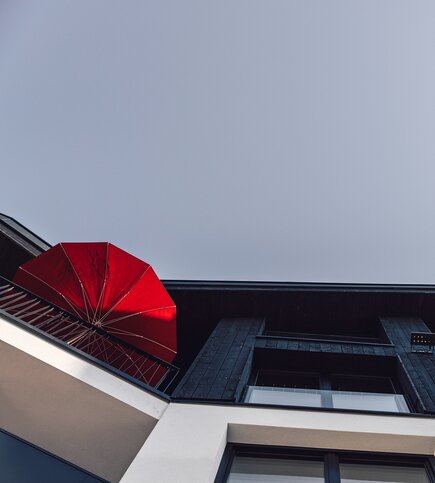 Ein roter Sonnenschirm auf einem Balkon vor einer dunklen Holzfassade und grauem Himmel.