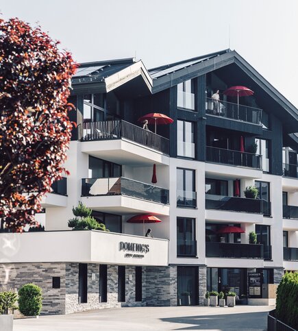 Außenansicht von Domenig's Luxury Apartments mit Balkonen und roten Sonnenschirmen.