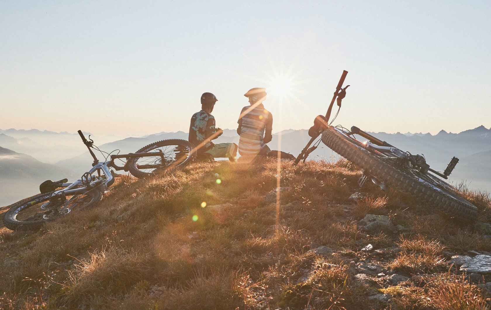 Zwei Personen stehen mit ihren Mountainbikes auf einem Gipfel im Sonnenlicht.