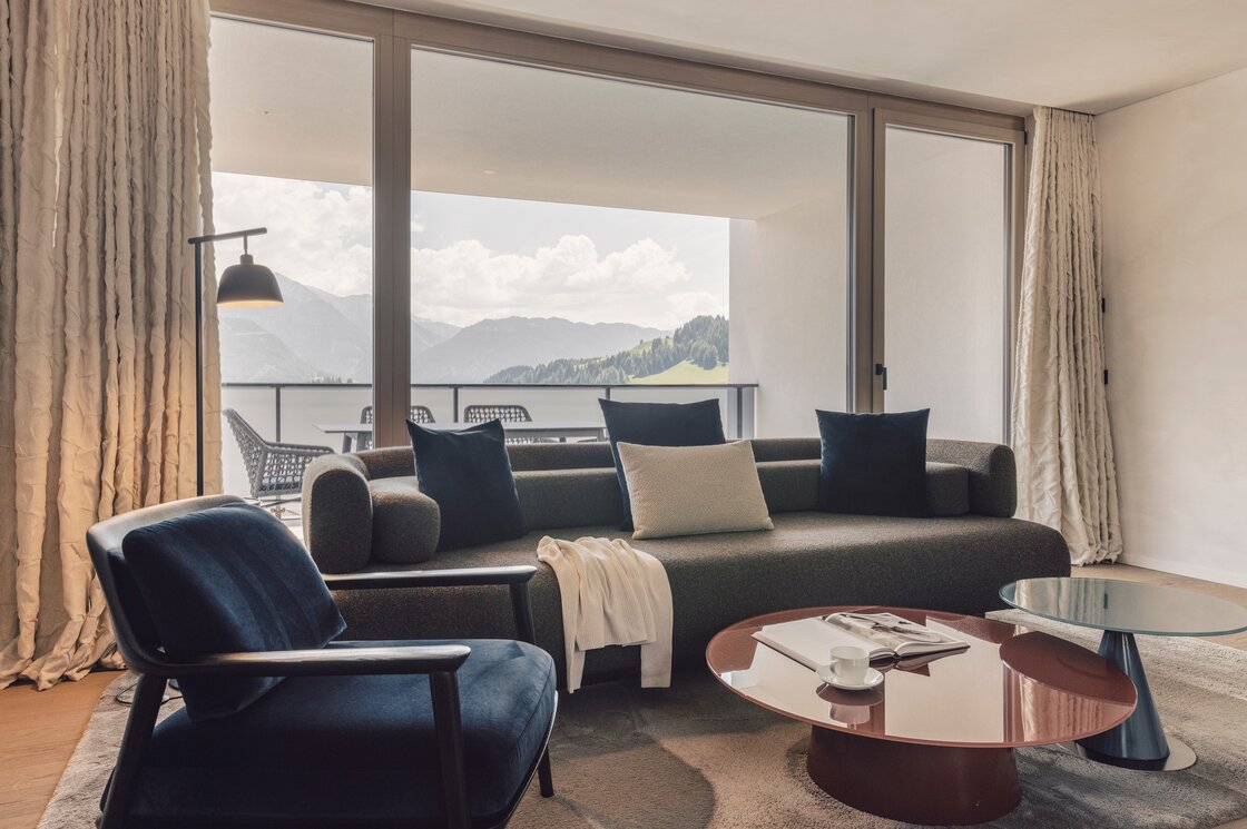 Wohnbereich eines Apartments mit Sofa, Sessel und großem Fenster zum Balkon mit Bergblick.
