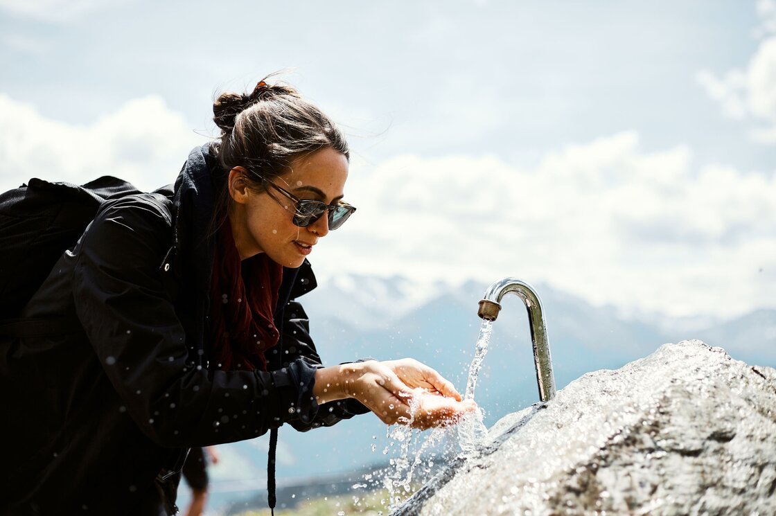 Eine Frau wäscht sich am Bergbrunnen die Hände, während im Hintergrund die Berge liegen.