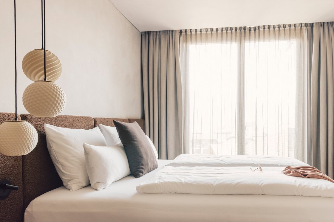 Ein modern eingerichtetes Schlafzimmer mit Doppelbett, großen Kissen und lichtdurchlässigen Vorhängen vor dem Fenster.