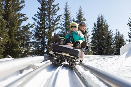 Zwei Kinder fahren auf einer Rodelbahn durch den Schnee, begleitet von einer erwachsenen Person.