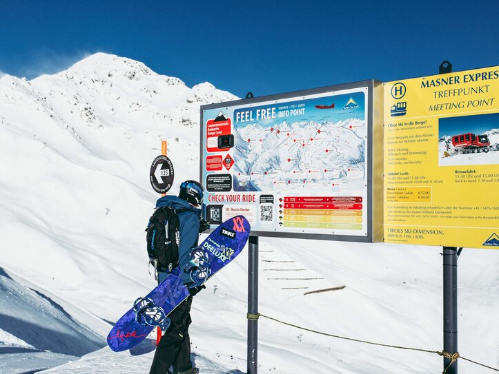 Ein Snowboarder steht vor Infotafeln im Schnee mit Bergkulisse in Serfaus-Fiss-Ladis.