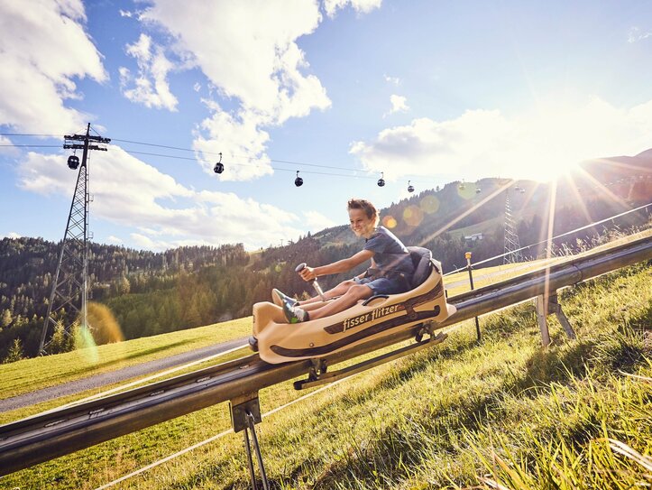 Sommerrodelbahn in Fiss mit Gondelbahn Ein Junge fährt auf der Sommerrodelbahn in Fiss bei sonnigem Wetter bergab, im Hintergrund verlaufen die Gondelkabinen.