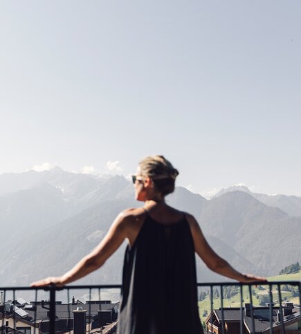 Eine Frau blickt vom Balkon auf die Berge und Dächer in Fiss.