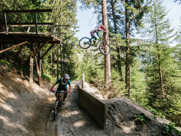 Mountainbiken in Serfaus-Fiss-Ladis im Waldtrail Zwei Mountainbiker fahren auf einem Waldtrail, einer springt über ein Hindernis neben einer hölzernen Plattform.