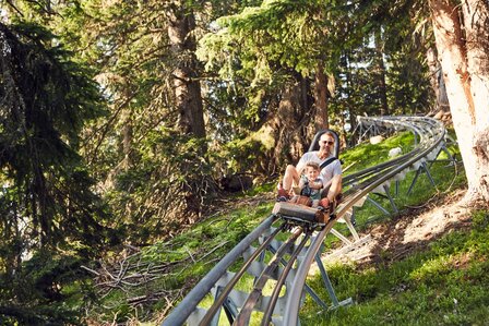 Ein Mann und ein Kind fahren mit der Sommerrodelbahn durch den Wald.