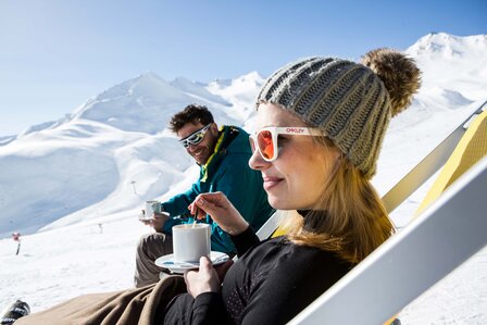 Zwei Personen sitzen mit Getränken im Schnee auf Liegestühlen vor einer verschneiten Bergkulisse.