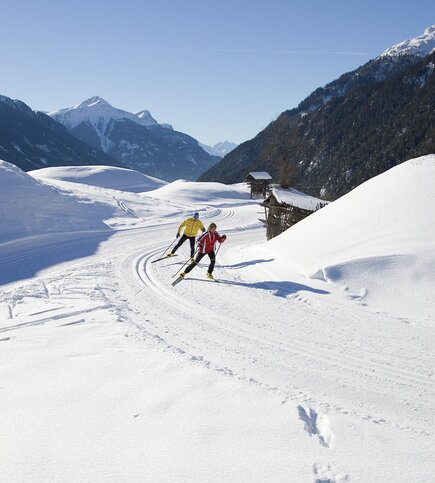 Zwei Langläufer sind auf einer gespurten Loipe in der verschneiten Berglandschaft unterwegs.