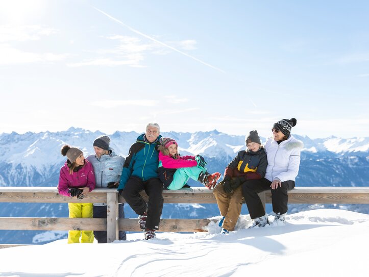 Eine Gruppe sitzt in Winterkleidung auf einer Holzbank mit Blick auf verschneite Berge.