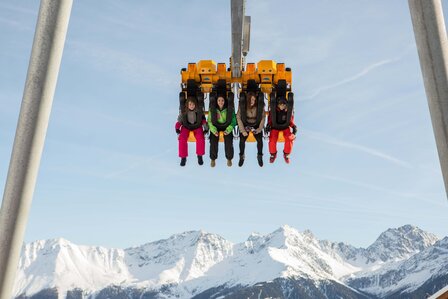 Vier Personen fahren in einem Vierer-Sessellift über eine winterliche Berglandschaft.