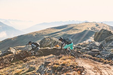 Zwei Mountainbiker fahren auf einem steinigen Bergpfad mit weiter Aussicht über die Tiroler Alpen.