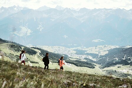 Drei Wanderer gehen über eine alpine Wiese mit Blick ins Tal und auf die Berge.