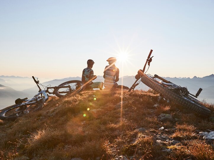 Mountainbiker mit Aussicht in Fiss Zwei Mountainbiker sitzen mit ihren Rädern auf einem Gipfel und blicken in die Berglandschaft im Sonnenlicht.