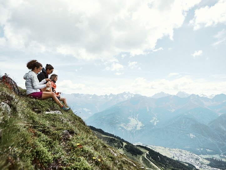 Drei Wanderinnen sitzen auf einer Bergwiese mit Blick ins weite Tal und auf die Alpen.