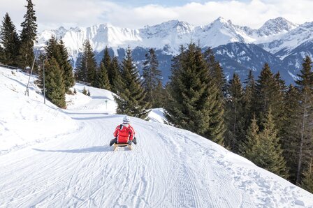 Eine Person rodelte auf einer verschneiten Piste mit Blick auf die Berge und Tannen.
