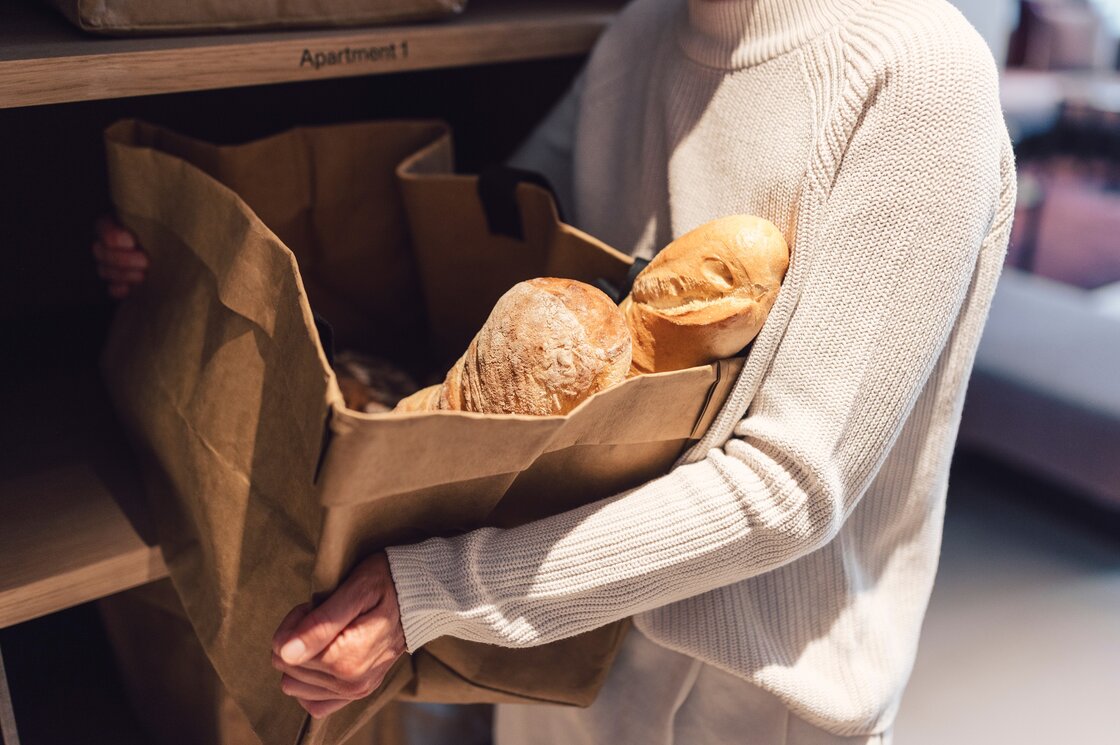 Eine Person hält einen Beutel mit frischen Brötchen vor einem Regal im Apartment.
