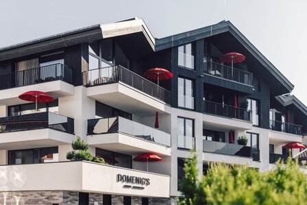 Außenansicht von Domenig's Luxury Apartments mit mehreren Balkonen und roten Sonnenschirmen.