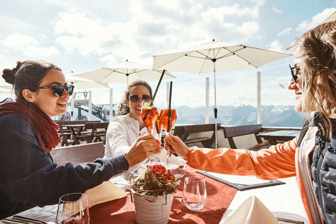 Drei Frauen stoßen auf einer Bergterrasse mit Aperitif und Blick auf die Tiroler Alpen an.