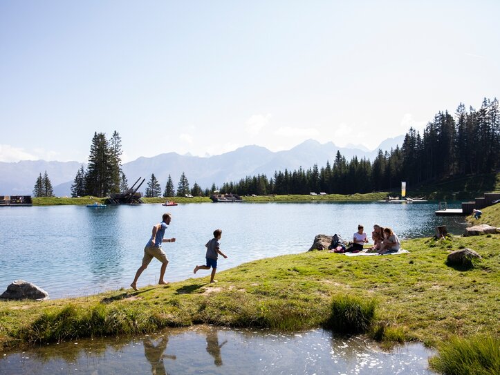 Menschen verbringen einen Sommertag am See mit Wiese, Bergen und Wald im Hintergrund.