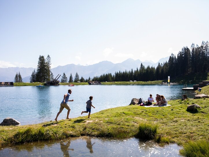 Sommerurlaub am See in Serfaus-Fiss-Ladis Menschen verbringen einen Sommertag am See mit Wiese, Bergen und Wald im Hintergrund.
