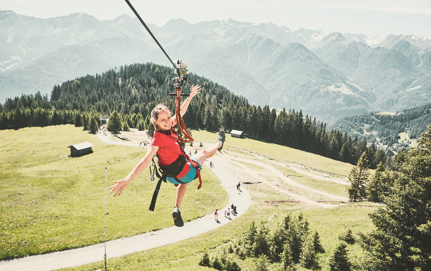 Eine Person schwebt an einer Seilrutsche über eine grüne Bergwiese mit Blick auf die Alpen.