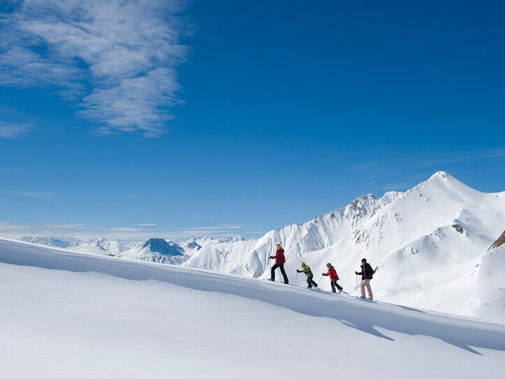 Vier Skitourengeher steigen auf einer verschneiten Bergkante vor den Gipfeln von Serfaus-Fiss-Ladis auf.