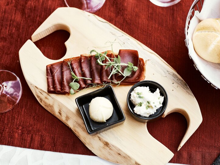 Ein Servierbrett mit aufgeschnittenem Trockenfleisch, Aufstrichen und Brötchen liegt auf einem roten Tisch.