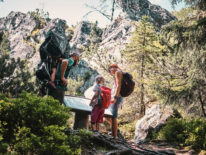 Wanderfamilie am Felsweg in Fiss Eine Familie mit Rucksack schaut auf einer Wanderung in Fiss auf eine Infotafel zwischen Felsen und Bäumen.
