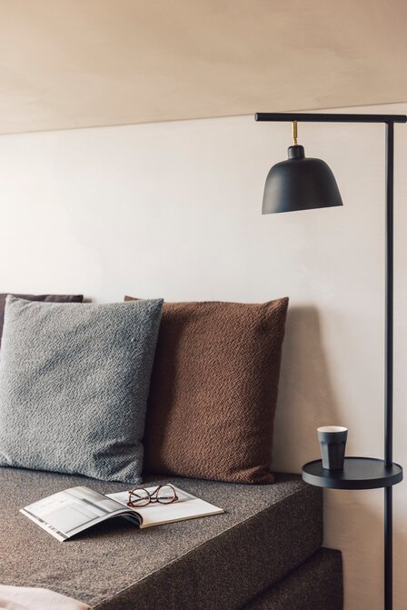 Ein Sofa mit Kissen steht neben einer schwarzen Leselampe; auf dem Sitz liegt ein offenes Magazin mit Brille.