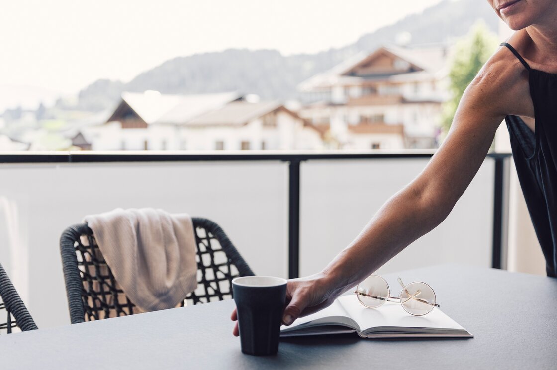 Eine Person stellt auf der Terrasse ein Getränk ab, mit Blick auf die Berge und Häuser im Hintergrund.