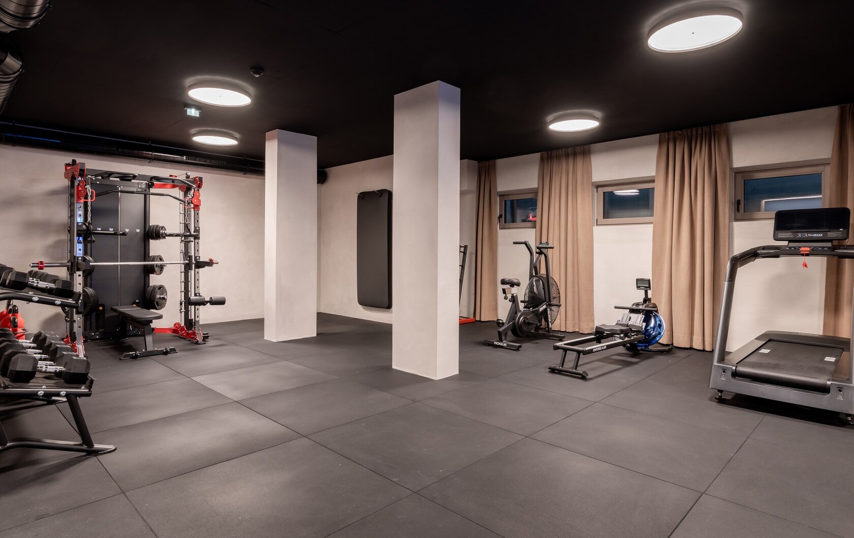 Der Fitnessraum zeigt Trainingsgeräte, Hanteln und ein Laufband in einem modernen Innenraum.