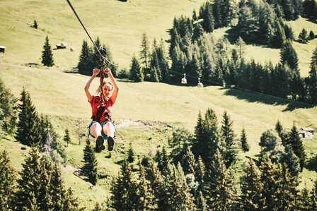 Eine Person fährt mit der Seilrutsche über eine grüne Bergwiese im Urlaubsgebiet Serfaus-Fiss-Ladis.