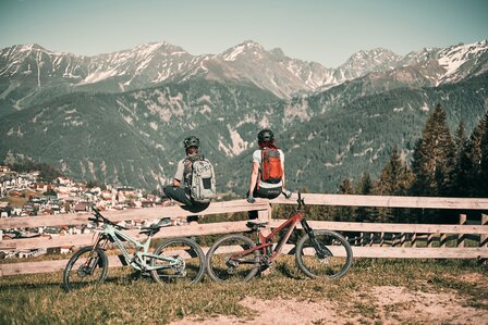 Zwei Mountainbiker sitzen an einer Holzbank mit Blick auf die Berge und das Tal bei Fiss.