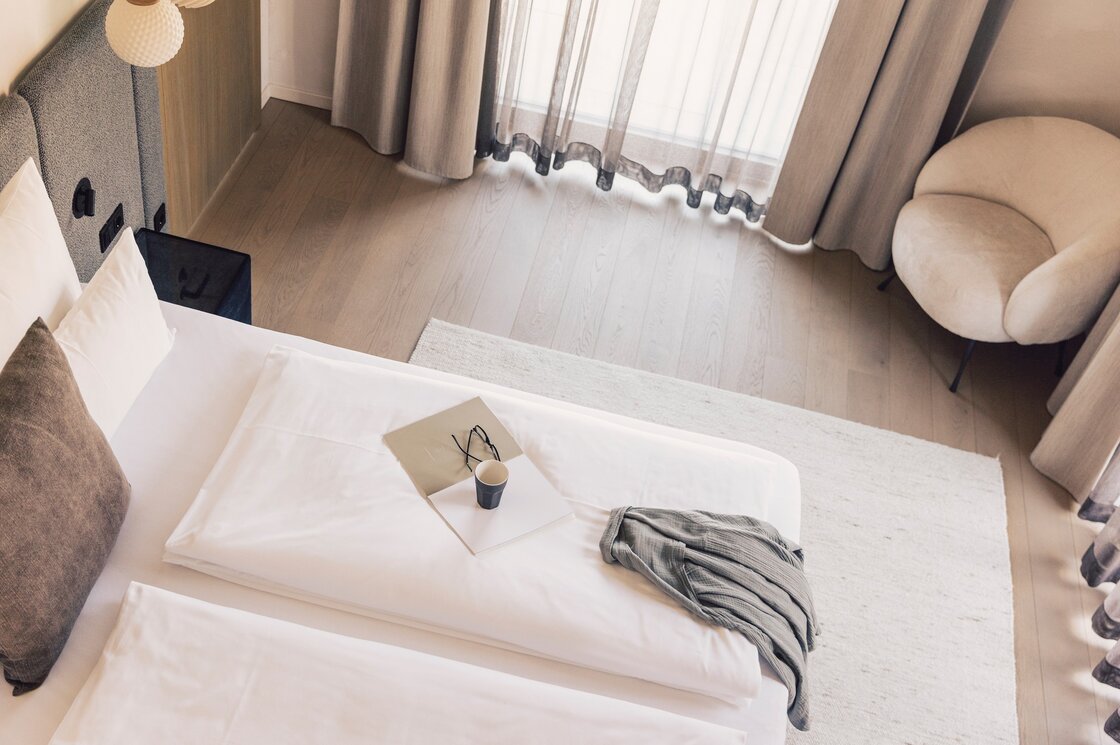 Ein helles Schlafzimmer mit Doppelbett, Sessel und großen Fenstern mit Vorhängen.