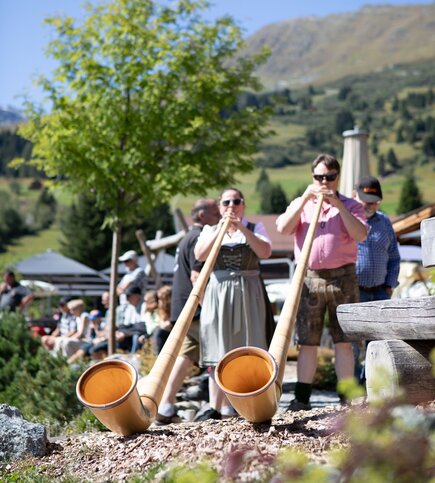 Mehrere Personen spielen Alphörner vor einer Bergkulisse im Sommer.