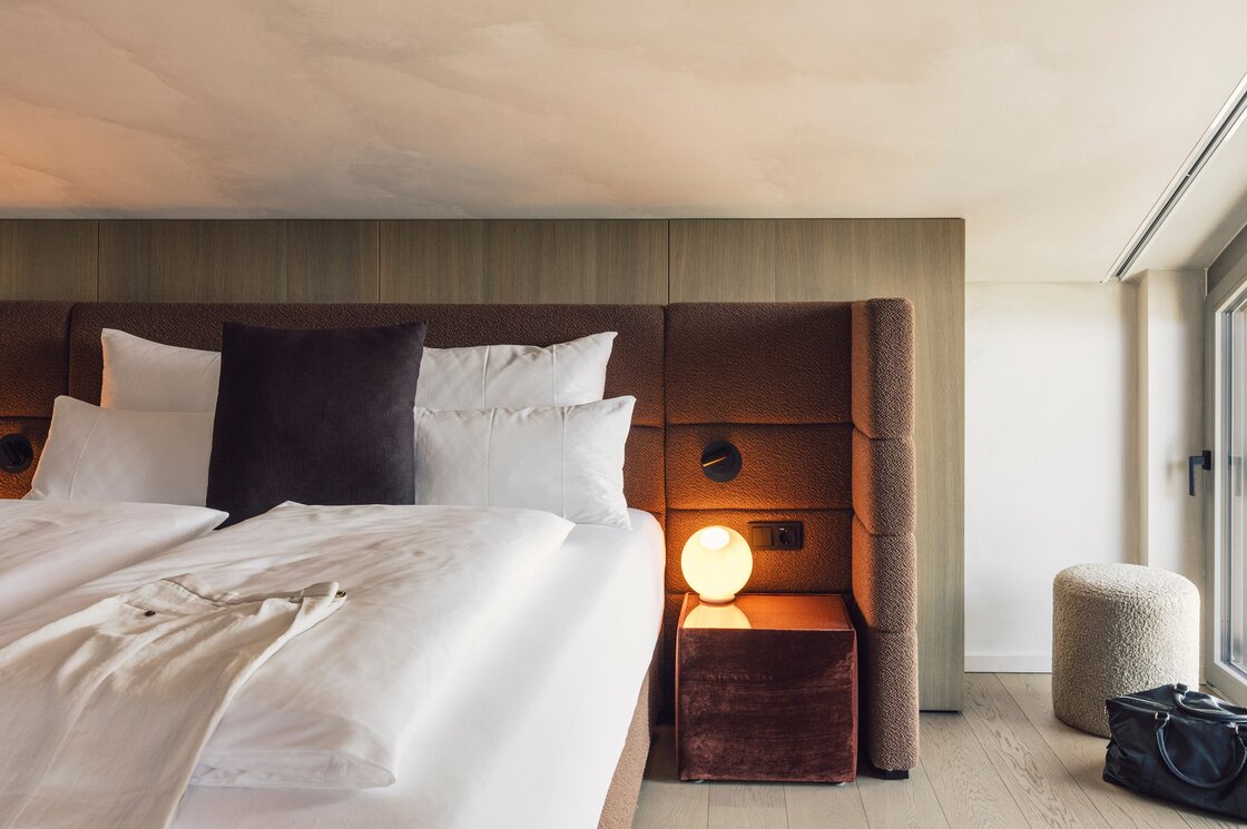 Ein modern eingerichtetes Schlafzimmer mit gepolstertem Kopfteil, großen Kissen und Fensterfront zur Seite.