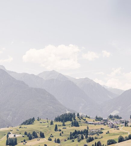 Eine weite Berglandschaft mit grünen Wiesen, verstreuten Bäumen und dem Ort Fiss im Tal.