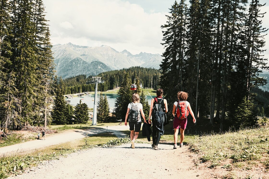 Drei Wanderer gehen auf einem Weg durch den Wald hinunter zum Bergsee mit Seilbahn in der Ferne.