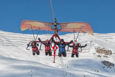 Vier Paragleiter fliegen über eine verschneite Berglandschaft unter blauem Himmel.