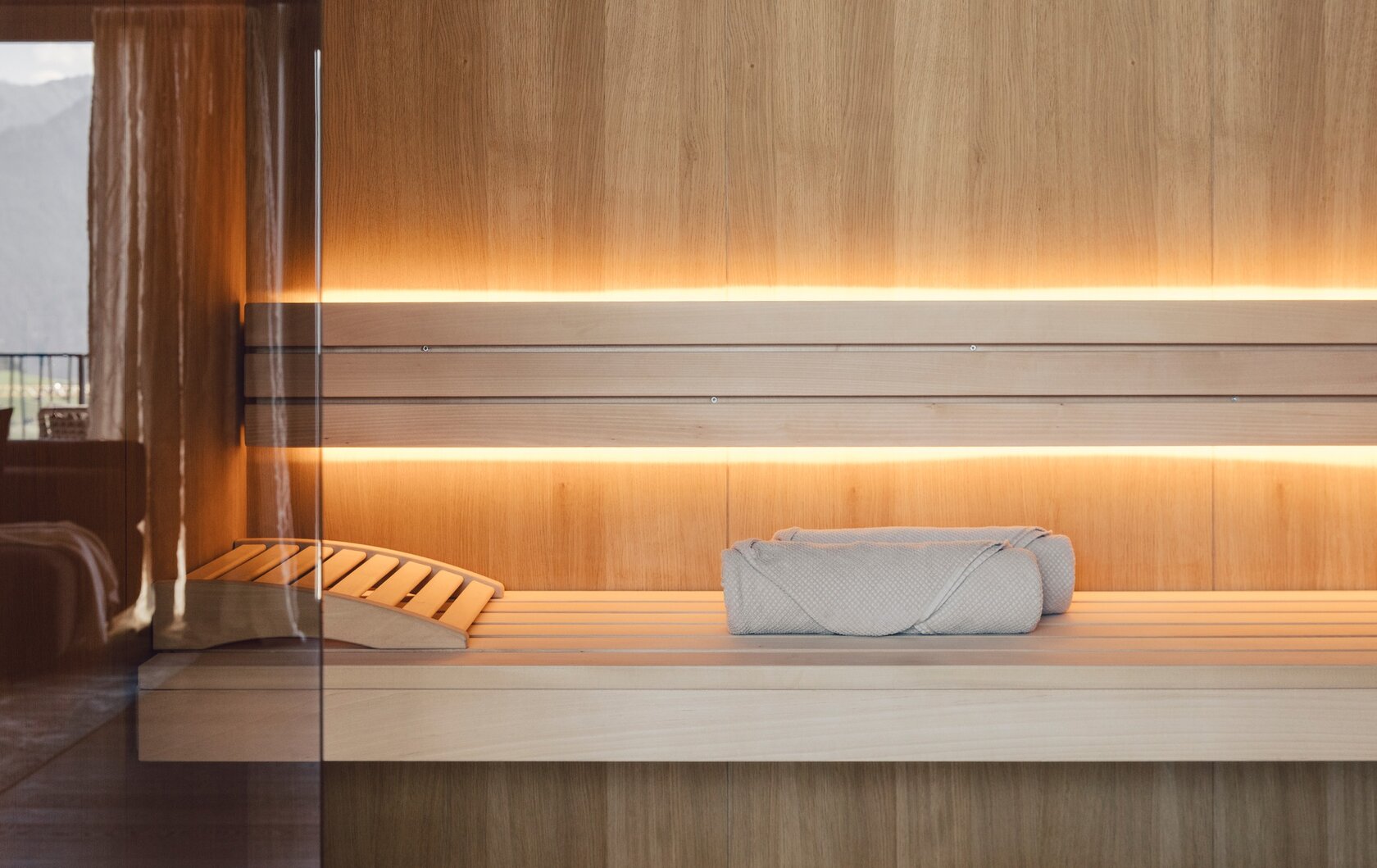 Eine Holzsauna mit Liegefläche und aufgerollten Handtüchern hinter Glas.