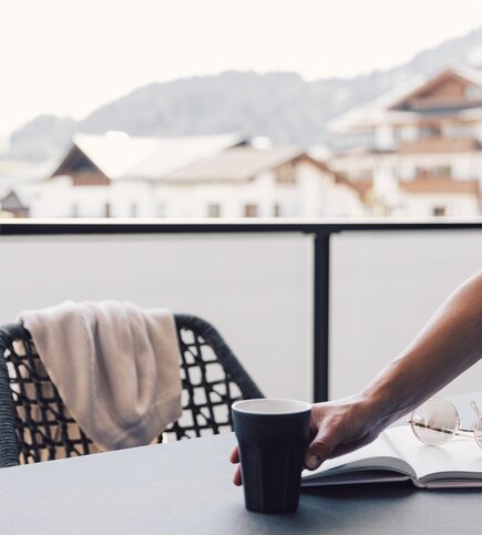 Auf dem Balkon steht ein Kaffeebecher neben einem aufgeschlagenen Buch, im Hintergrund sind Berge und Häuser zu sehen.