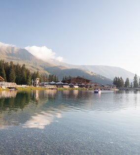 Ein ruhiger Bergsee mit Holzhütten, Wäldern und Bergen im Hintergrund.