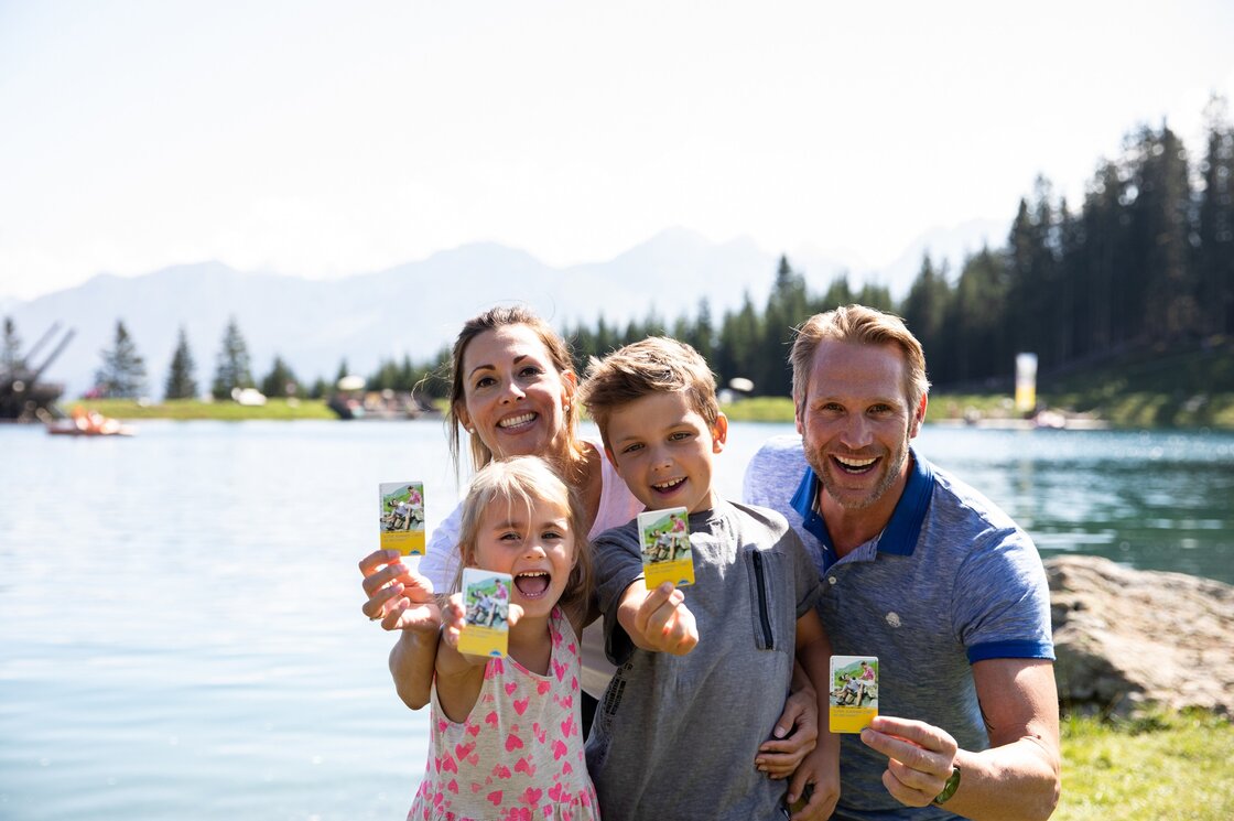 Eine Familie hält am Bergsee mehrere Karten in die Kamera, im Hintergrund liegen Wälder und Berge.