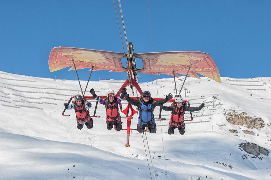 Vier Wintersportler sitzen auf einem Sessellift über einer verschneiten Berglandschaft.